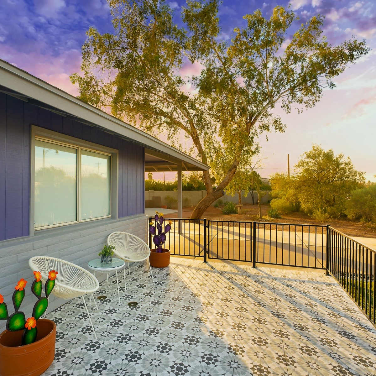 A colorful patio with white chairs, potted cacti, and decorative plants, alongside a modern home under a sunset sky.