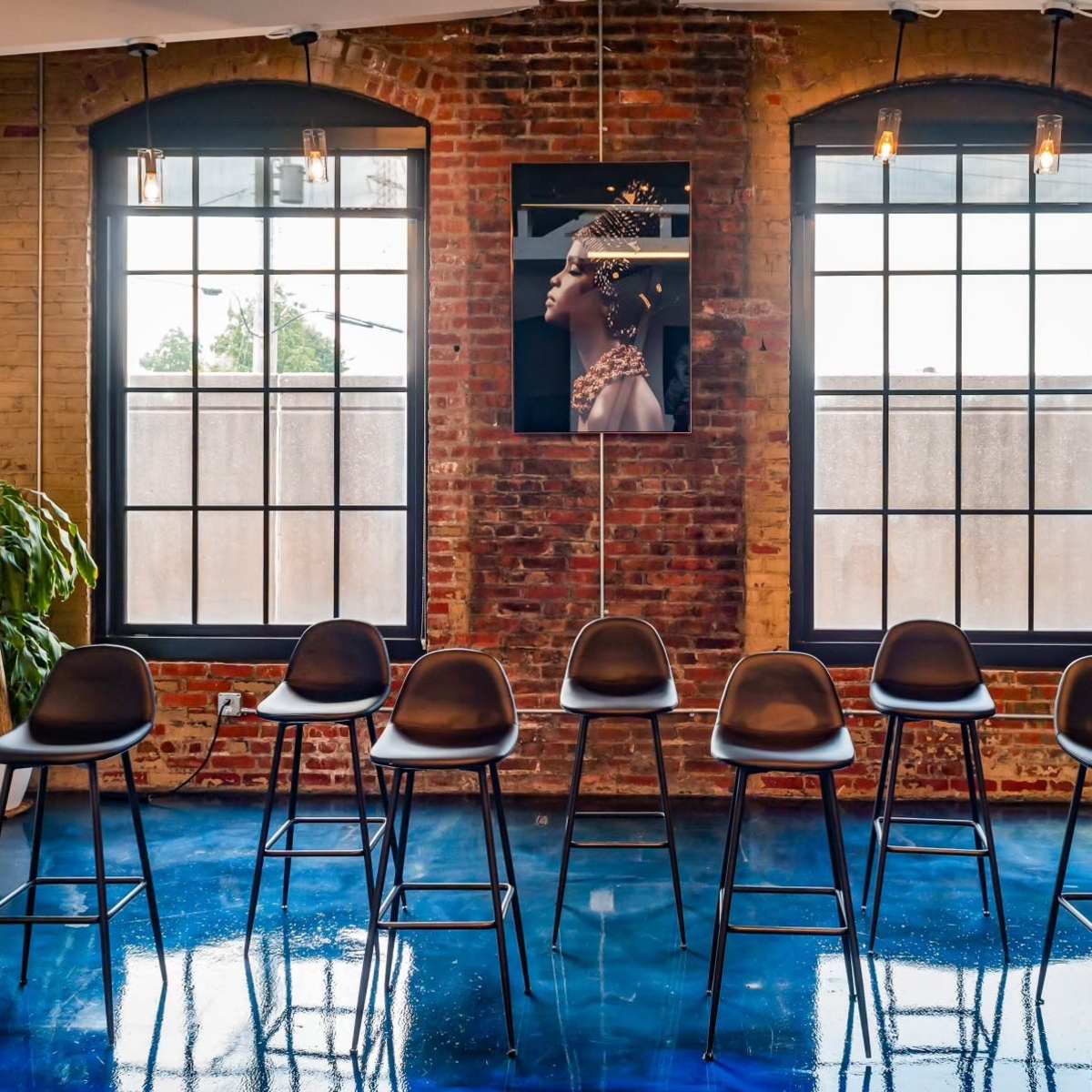 A line of black bar stools is arranged in front of large windows with a brick wall and a framed portrait hanging between them.
