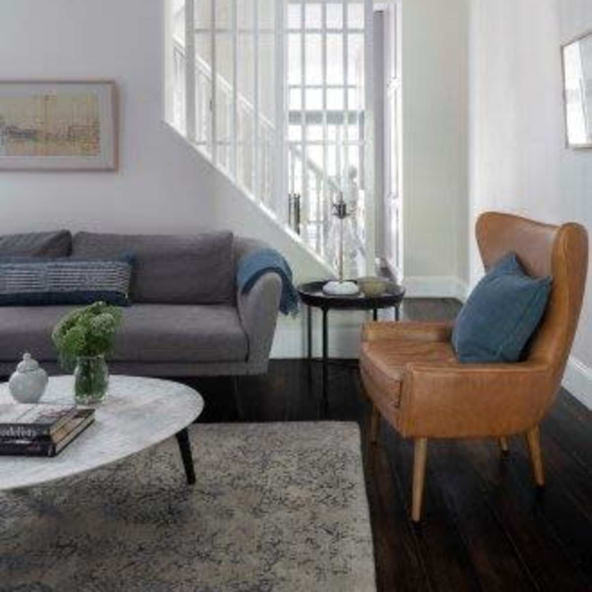 A modern living room features a gray sofa, a brown armchair, a marble coffee table, and a staircase in the background.