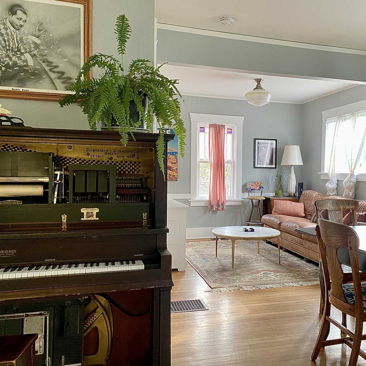A cozy living space with a vintage piano, a dining table with chairs, a leather couch, and various plants scattered throughout the room.