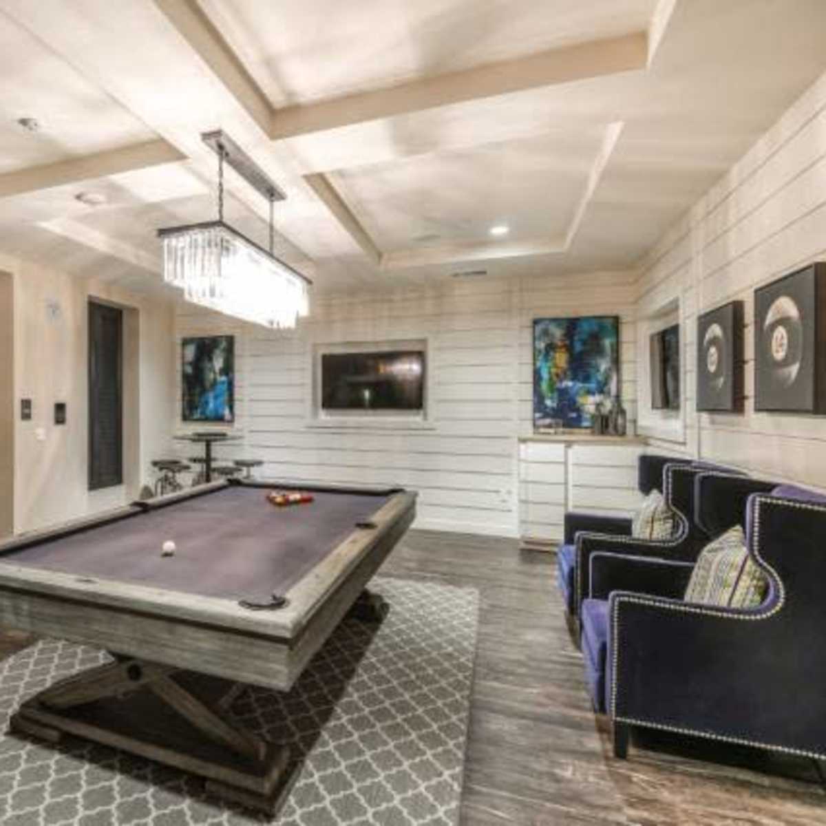 A game room with a pool table, two armchairs, and a modern chandelier, set against a backdrop of paneled walls and artwork.