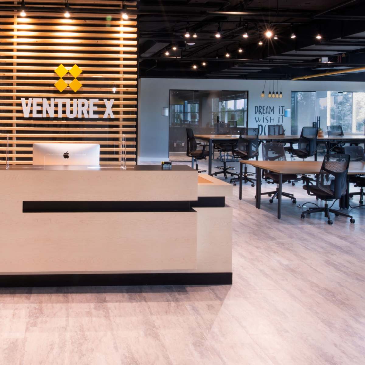 A modern reception area with a sleek wooden desk, a large sign reading "VENTURE X," and a workspace featuring multiple office tables and chairs.
