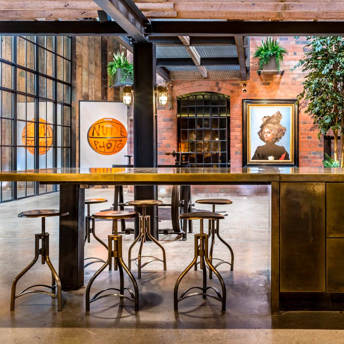 A modern industrial kitchen area with a large metallic island, wooden stools, and a vibrant artwork on the wall.