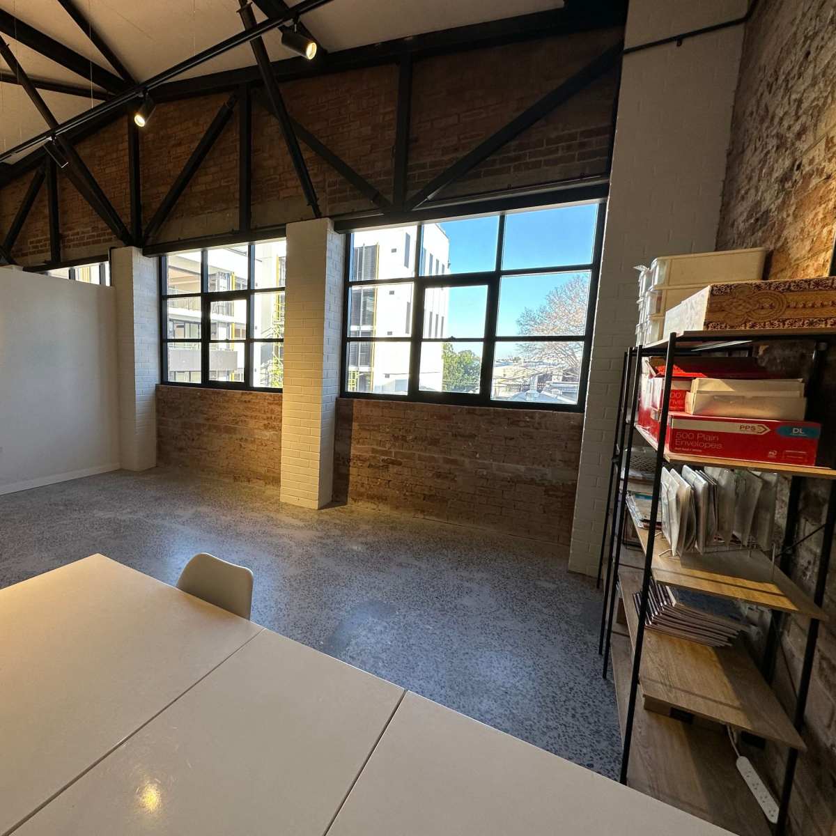 An empty room with exposed brick walls, large windows, and minimal furniture including a table and chairs.