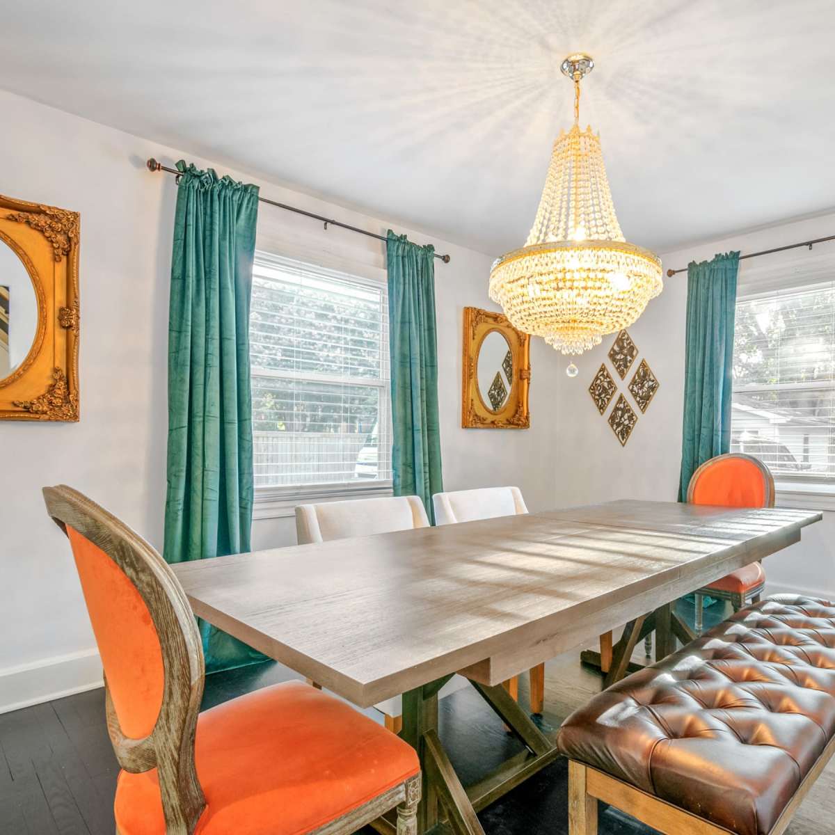A dining room features a long wooden table surrounded by chairs, with a chandelier and mirrors on the walls.