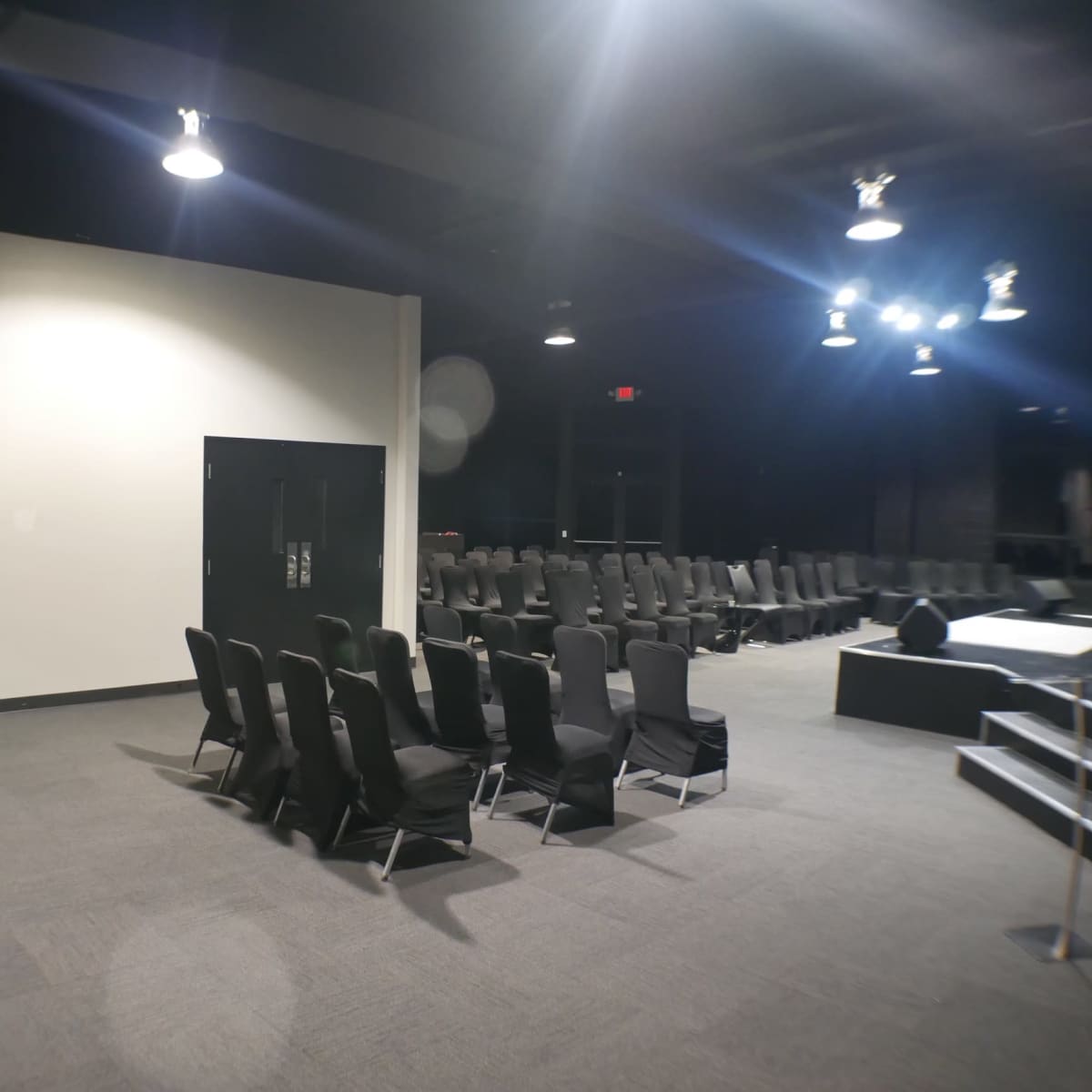 A spacious room features rows of black chairs facing a stage, with a door and a fire extinguisher visible on adjacent walls.