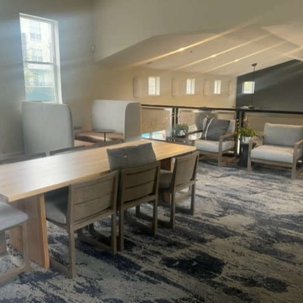 A spacious semi-enclosed lounge area featuring a long wooden table surrounded by chairs, cozy seating along the walls, and large windows allowing natural light to enter.