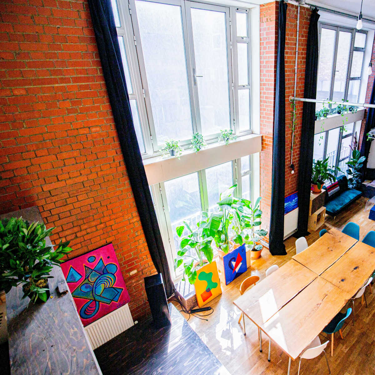 A spacious room with large windows, a long wooden table, colorful artwork on the walls, and a seating area filled with plants.