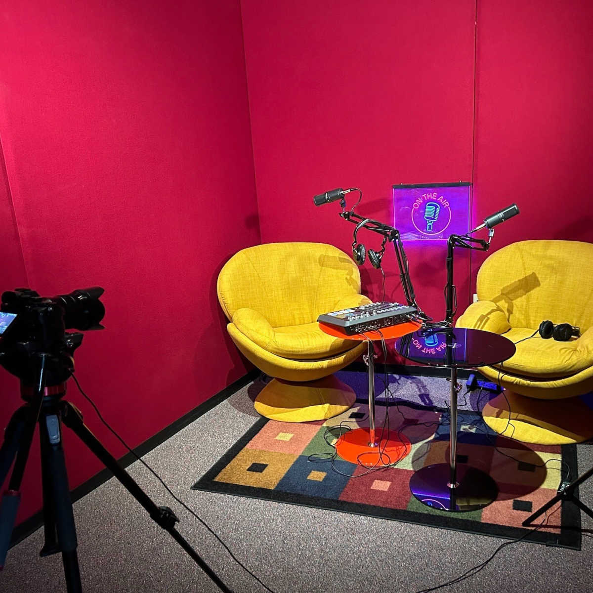 A recording studio with two yellow chairs, a microphone setup, a small table with audio equipment, and a camera pointing towards the chairs.