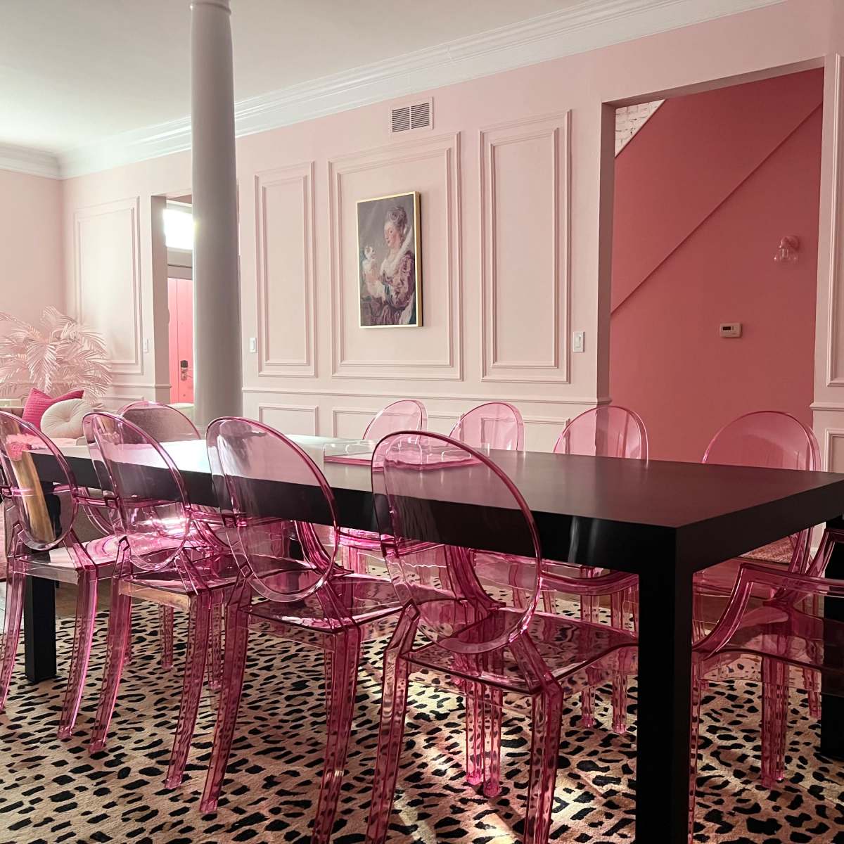 A rectangular black dining table surrounded by transparent pink chairs sits on a leopard-print rug in a room with pink walls and wainscoting.