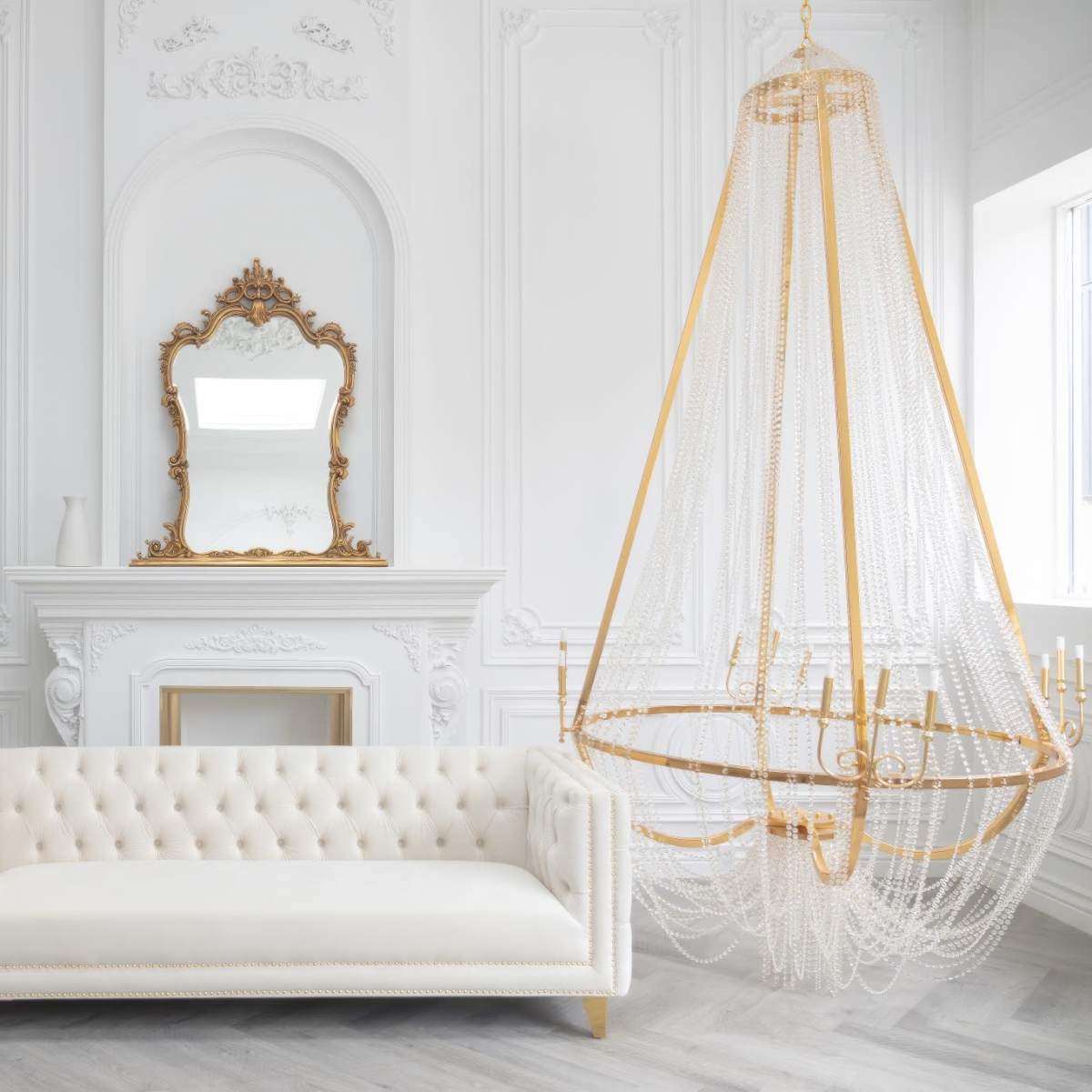 A luxurious white tufted sofa facing a decorative fireplace, with an ornate mirror above it and an elegant chandelier hanging from the ceiling.