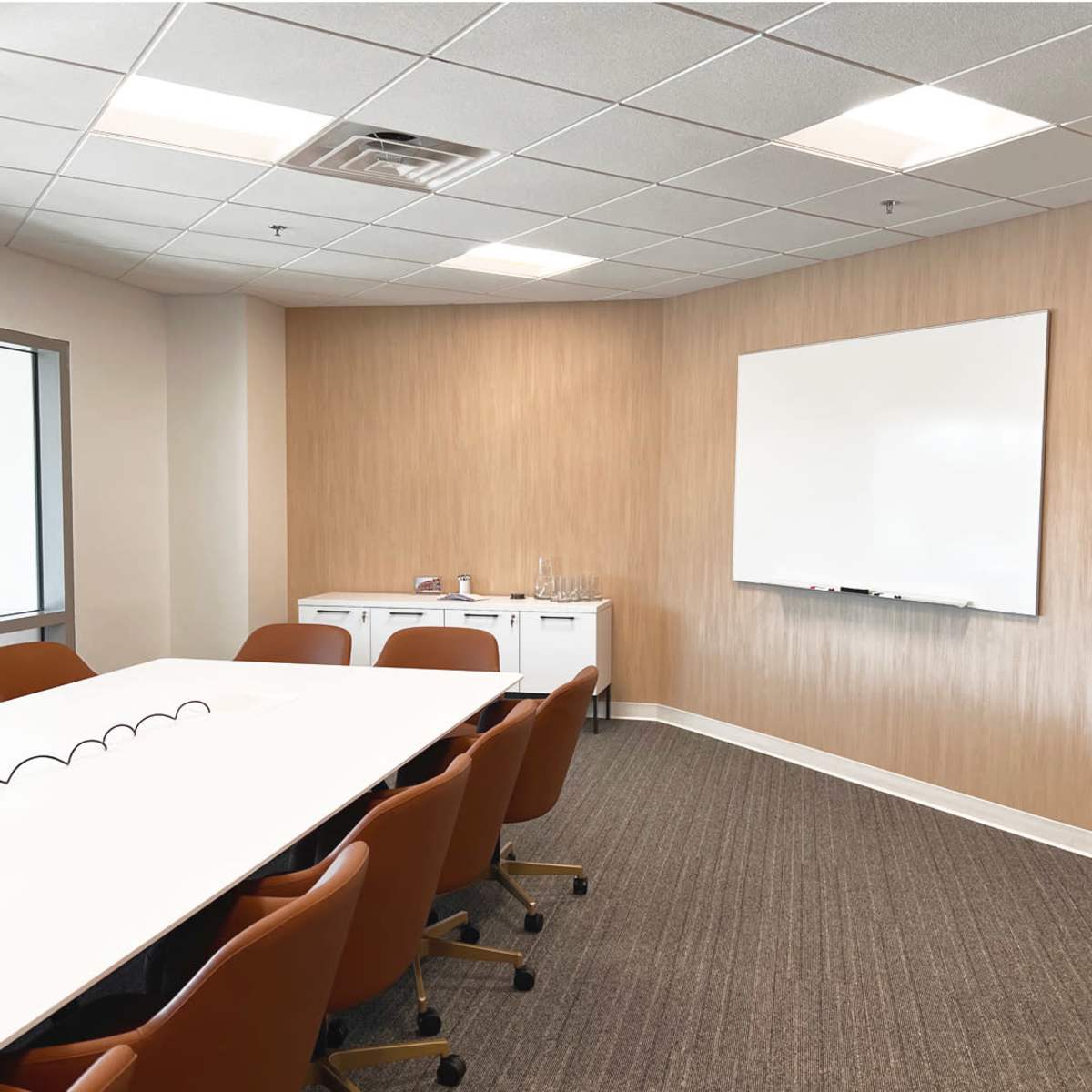 A conference room features a whiteboard, a long table surrounded by brown chairs, and a light-colored wall with a textured finish.
