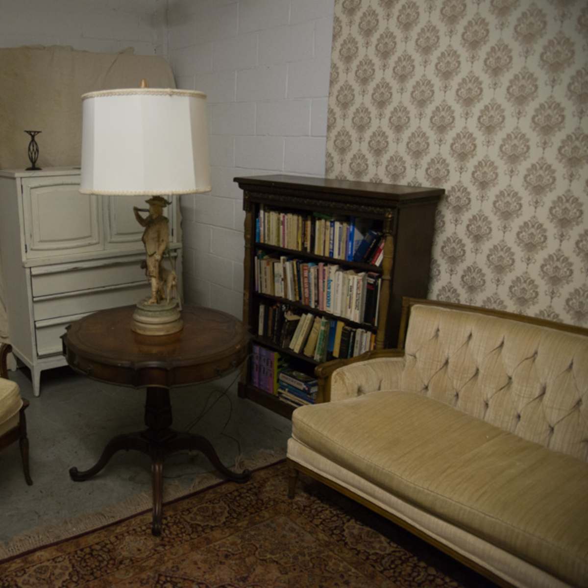 A small sitting area with two beige upholstered chairs, a round wooden table, and a lamp, set against a wall with wallpaper and a bookshelf filled with books.