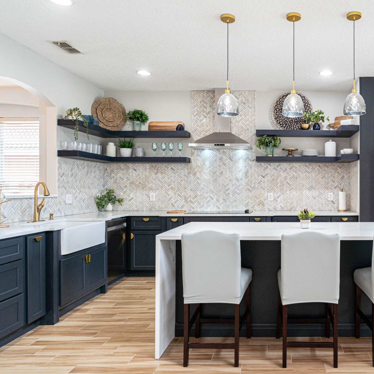 A modern kitchen with dark cabinetry, a white countertop, and three upholstered bar stools at an island, featuring a patterned backsplash and various plants for decoration.