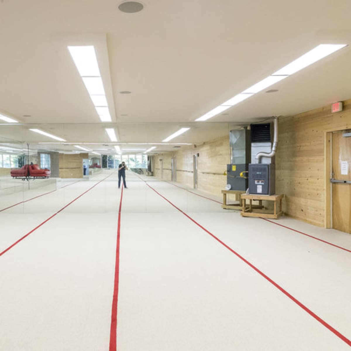 A spacious gymnasium features a mirrored wall, red lines marking the gym floor, and a person standing at the center.
