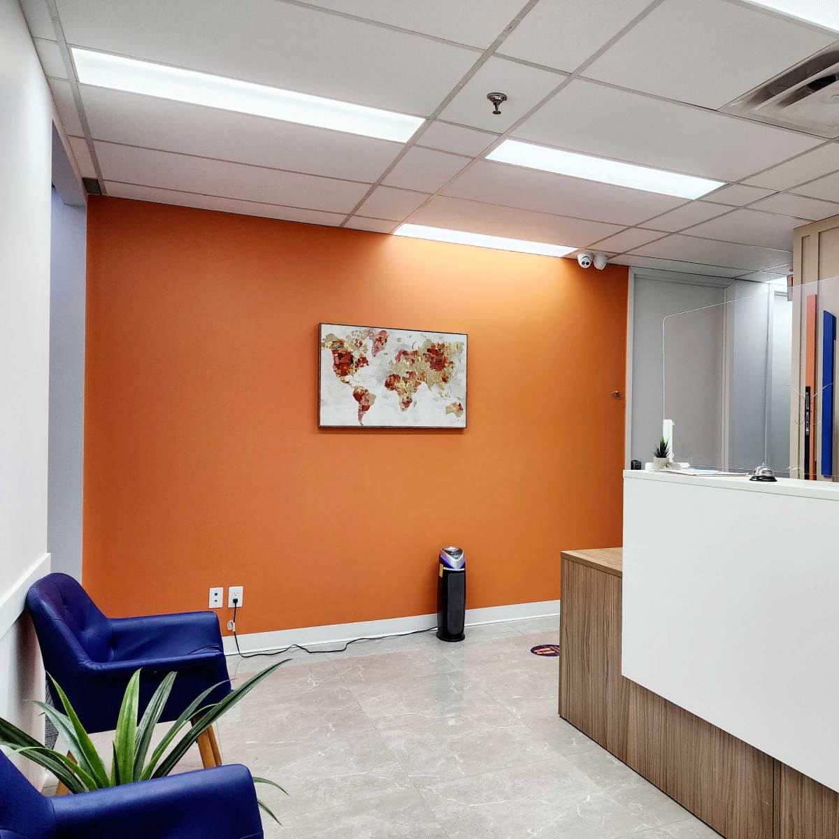 A modern office reception area with orange walls, a white desk with a protective divider, and blue chairs.