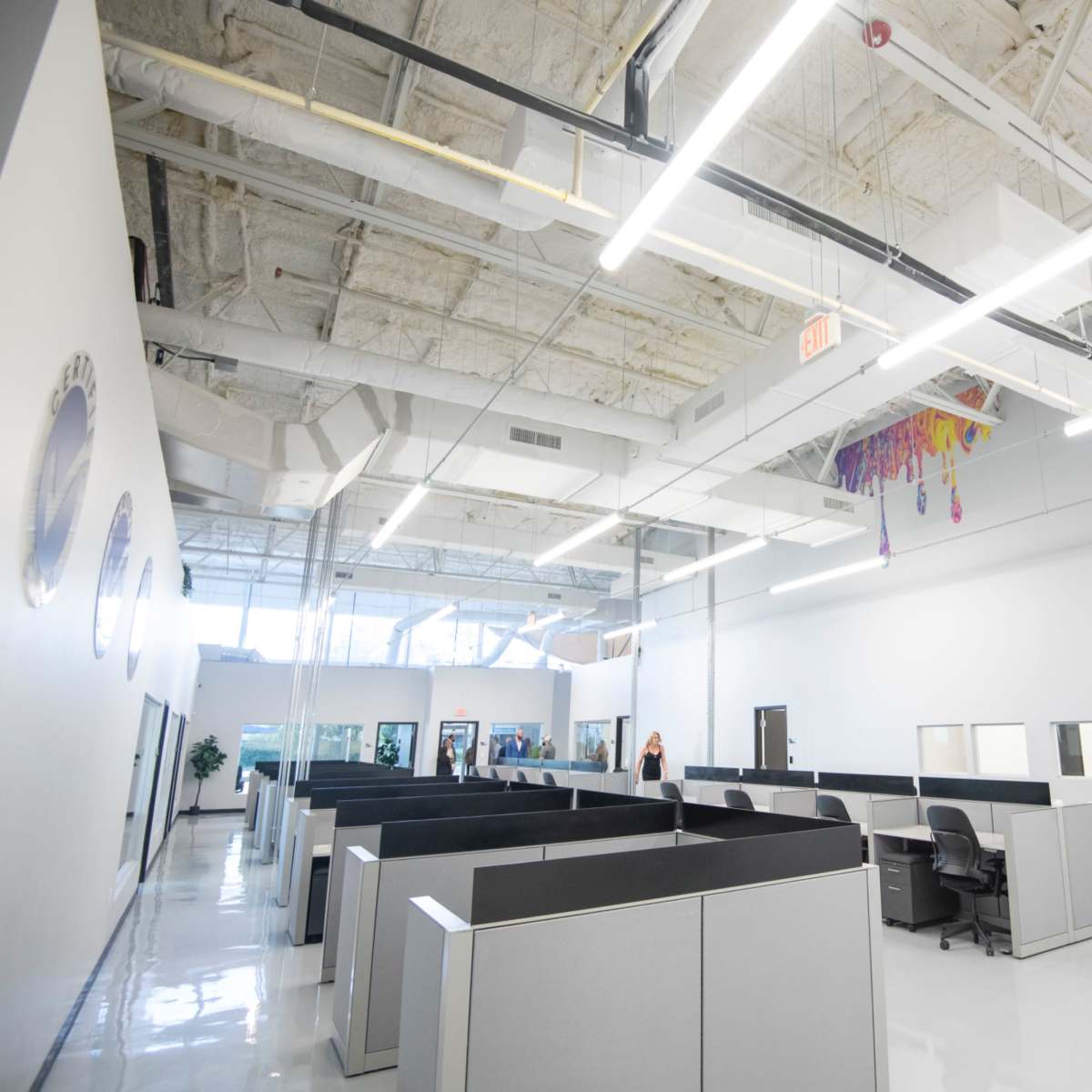A spacious, modern office with gray cubicles arranged in a row under bright overhead lighting.