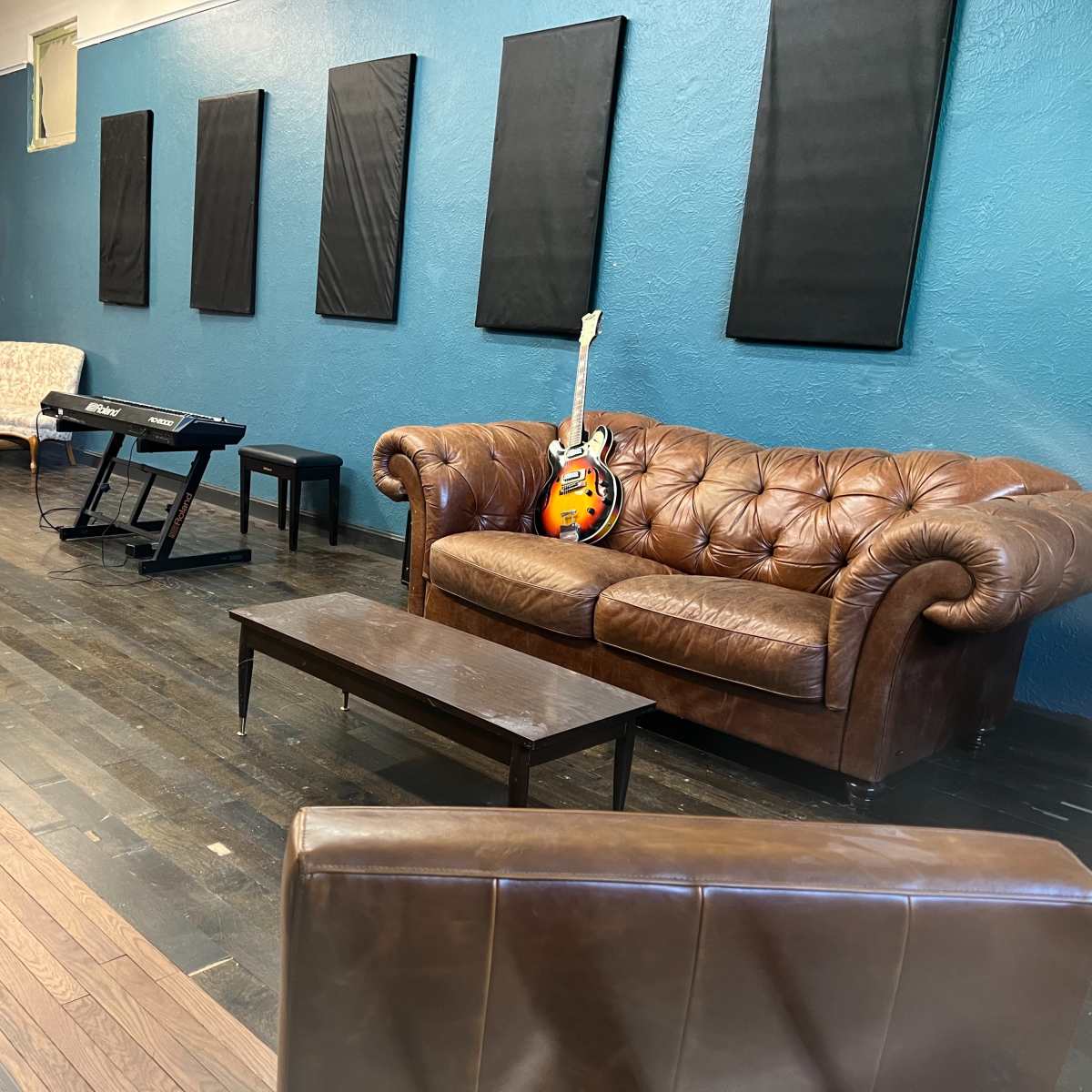 A room with a brown leather couch, a small coffee table, a keyboard on a stand, and an electric guitar leaning against the couch, all set against a blue wall with soundproofing panels.