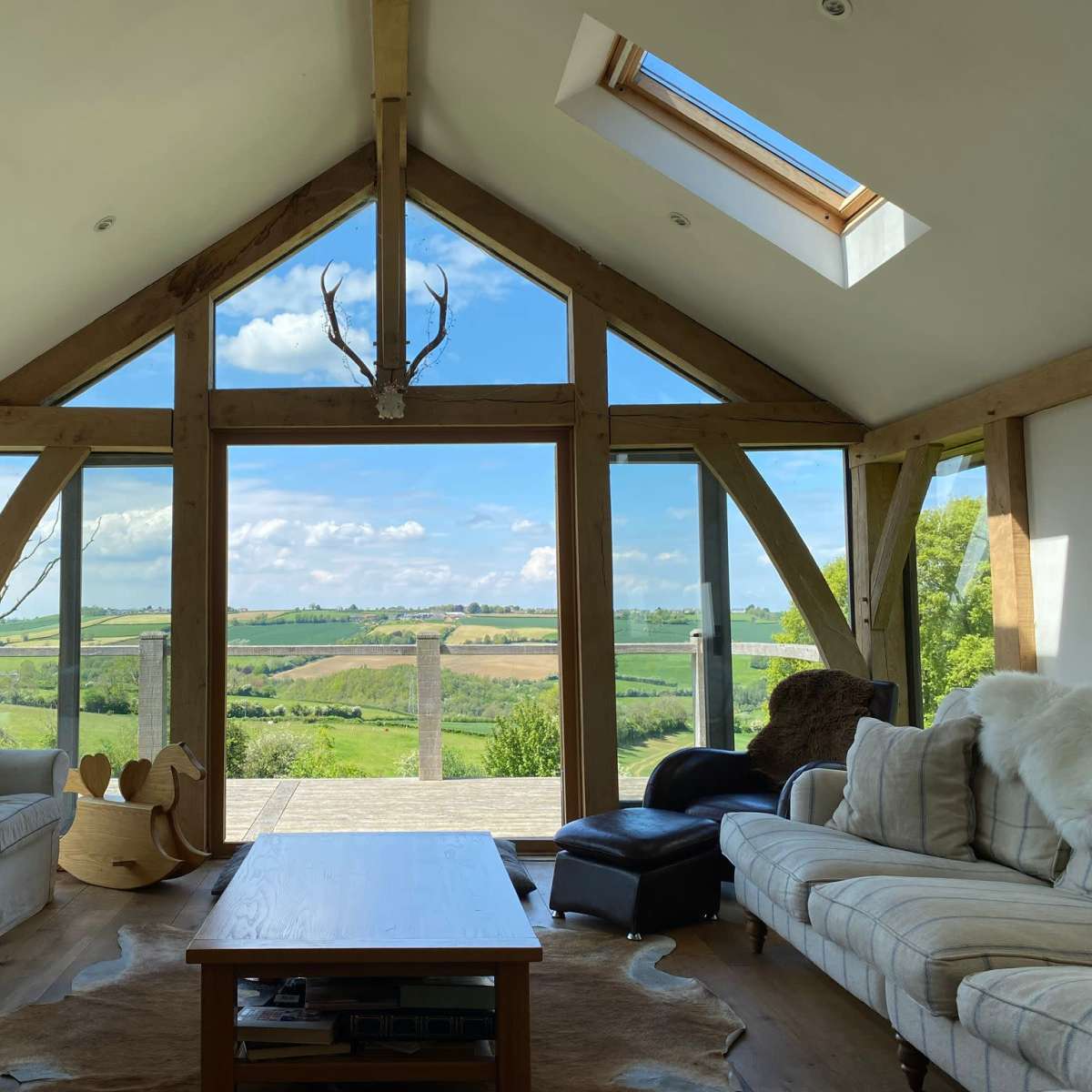 A modern living room with large windows overlooking a green landscape and wooden beams supporting the ceiling.