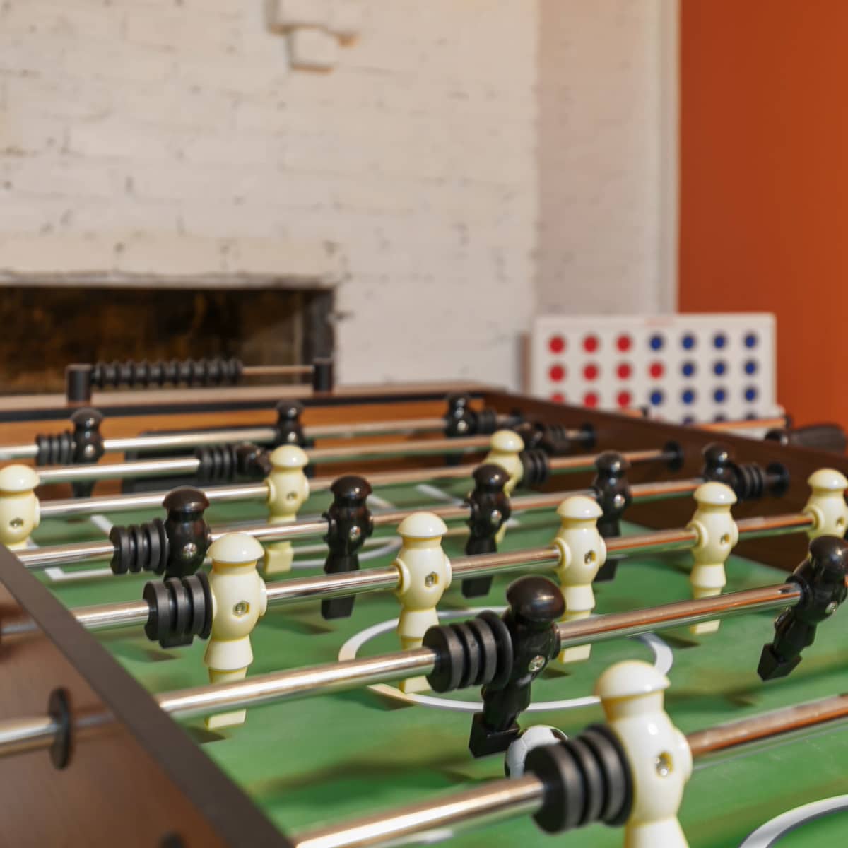 A foosball table with black and white figures is positioned in a room with an orange wall and a faint background of a Connect Four game.
