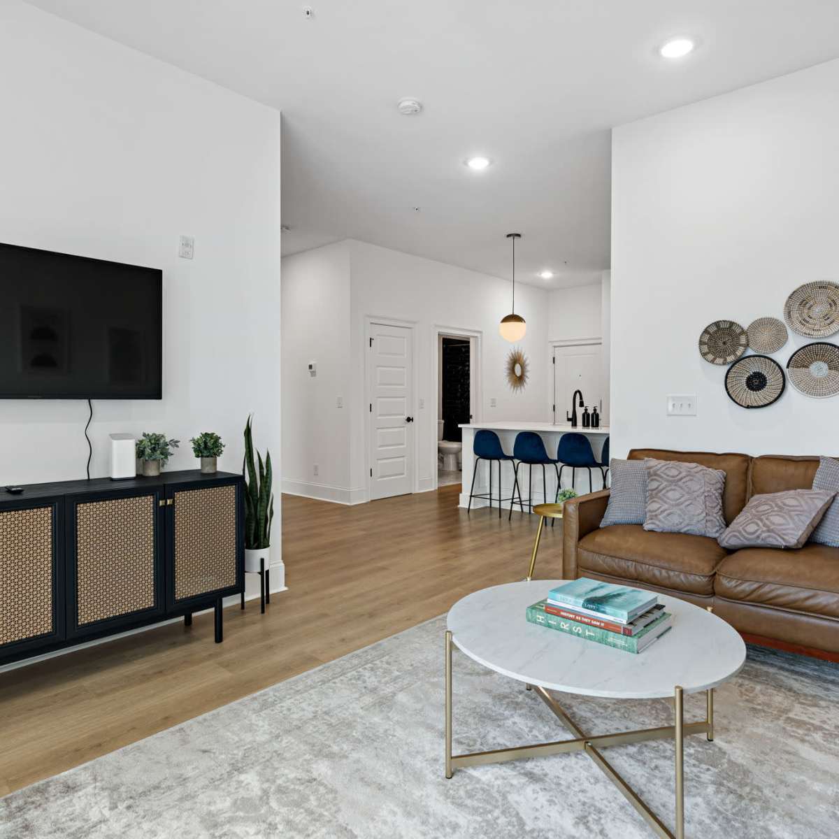 A modern living room with a brown sectional sofa, a circular coffee table, a wall-mounted TV, and a dining area in the background featuring bar stools and decorative plates on the wall.