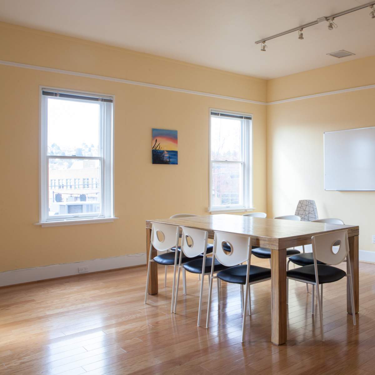 A bright, empty conference room featuring a wooden table surrounded by chairs, two windows, a whiteboard, and a plant in the corner.