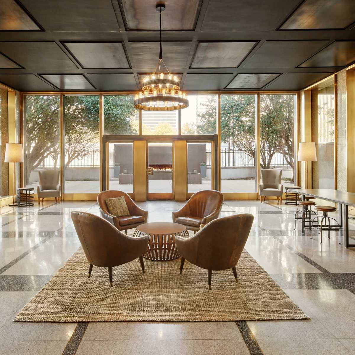 A modern lobby features two leather armchairs facing a round coffee table on a woven rug, with large windows providing an outside view and a chandelier hanging overhead.