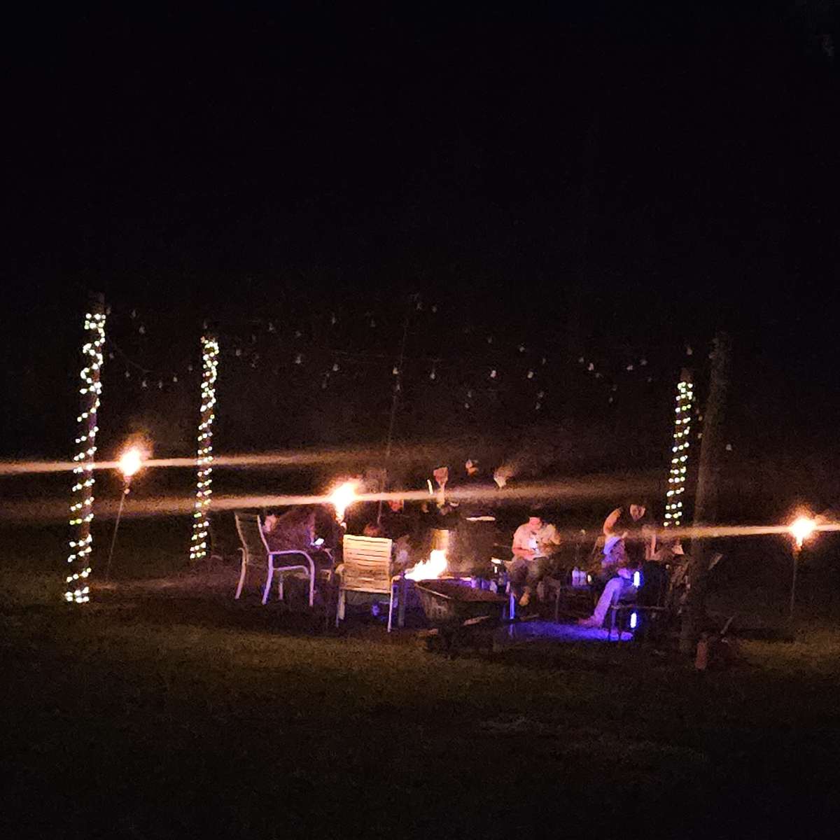 A group of people gathers around a fire pit, surrounded by glowing torches and string lights in a dark outdoor setting.