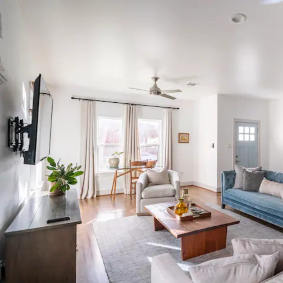 The living room features a blue velvet couch, a light-colored sofa, a coffee table, and a window with sheer curtains, complemented by a small dining table and light wood flooring.