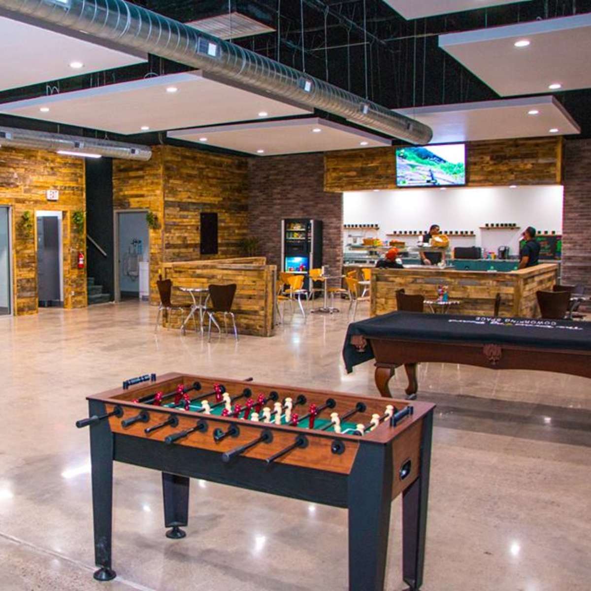 A spacious indoor recreational area featuring a foosball table, seating, a bar, and a screen displaying information.