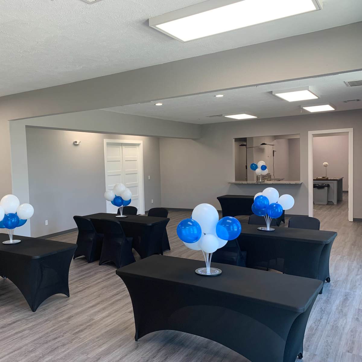 A rental space with several tables covered in black cloth, each adorned with blue and white balloons, set up for an event.