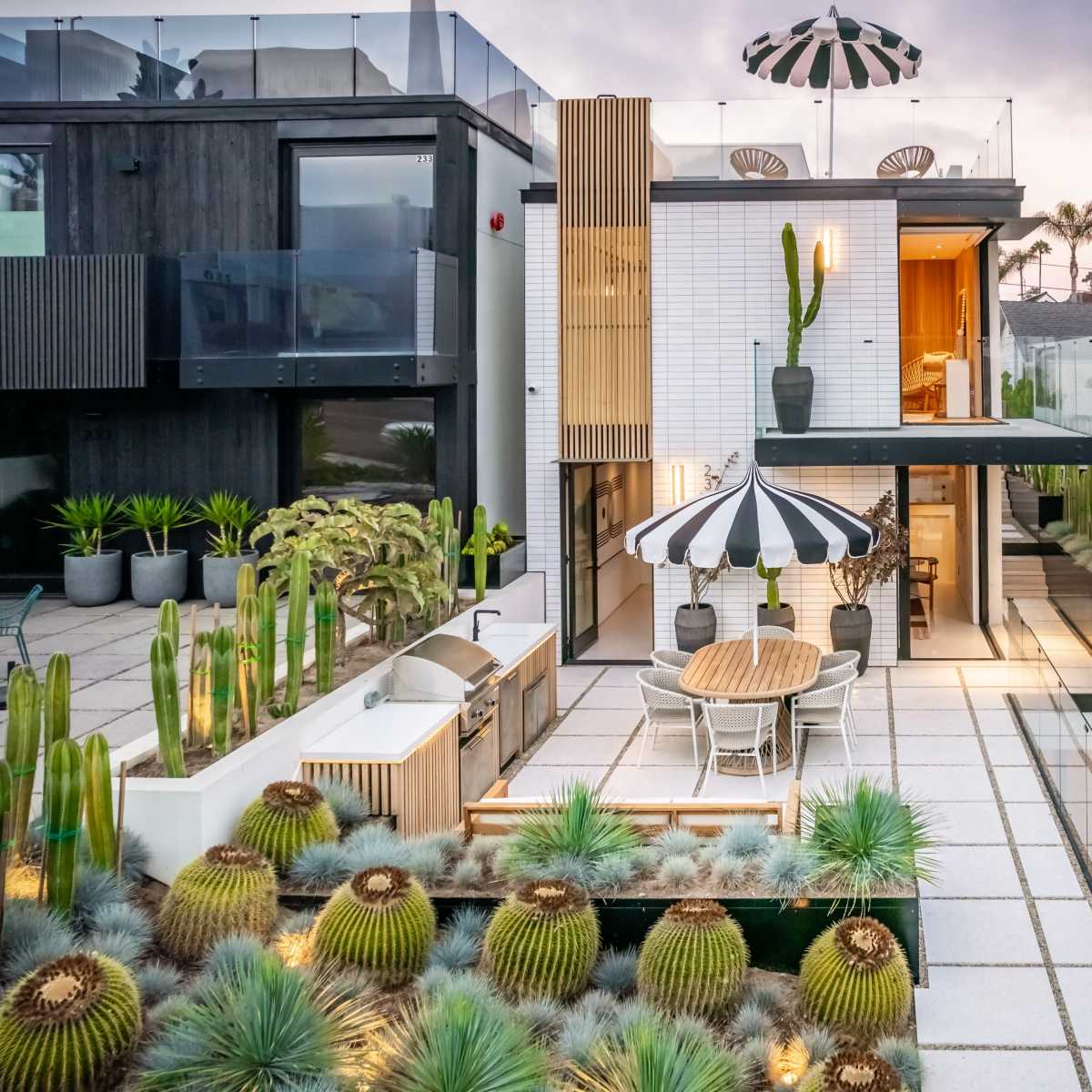 A modern house with a rooftop terrace featuring a striped umbrella, surrounded by a landscaped garden filled with cacti and a dining area.