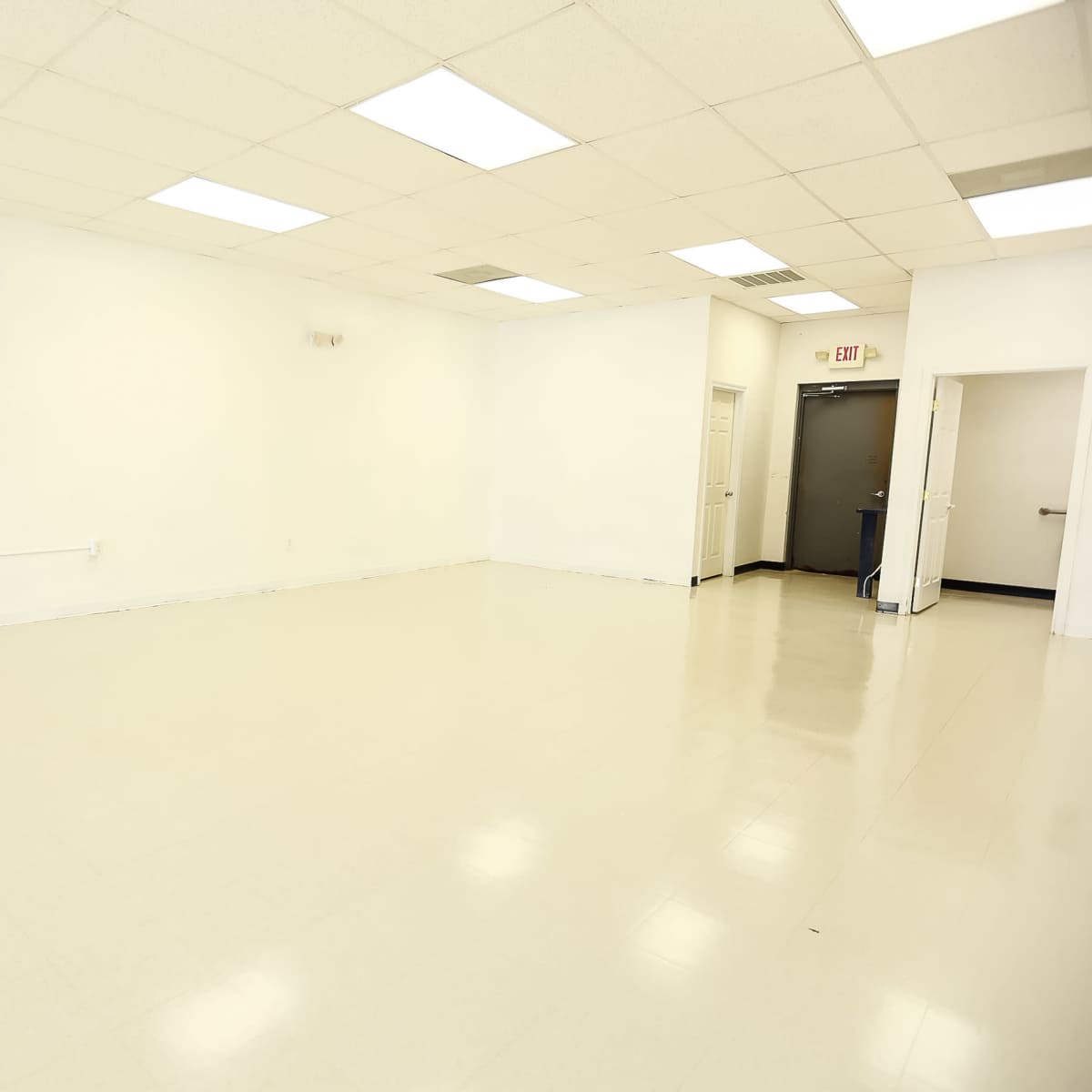 A large, empty room with white walls and a smooth, light-colored floor, featuring fluorescent ceiling lights.