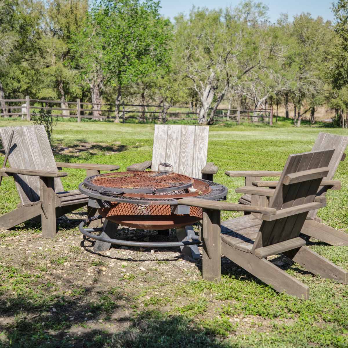 A circular fire pit surrounded by four wooden chairs sits in a grassy area with trees and a fence in the background.