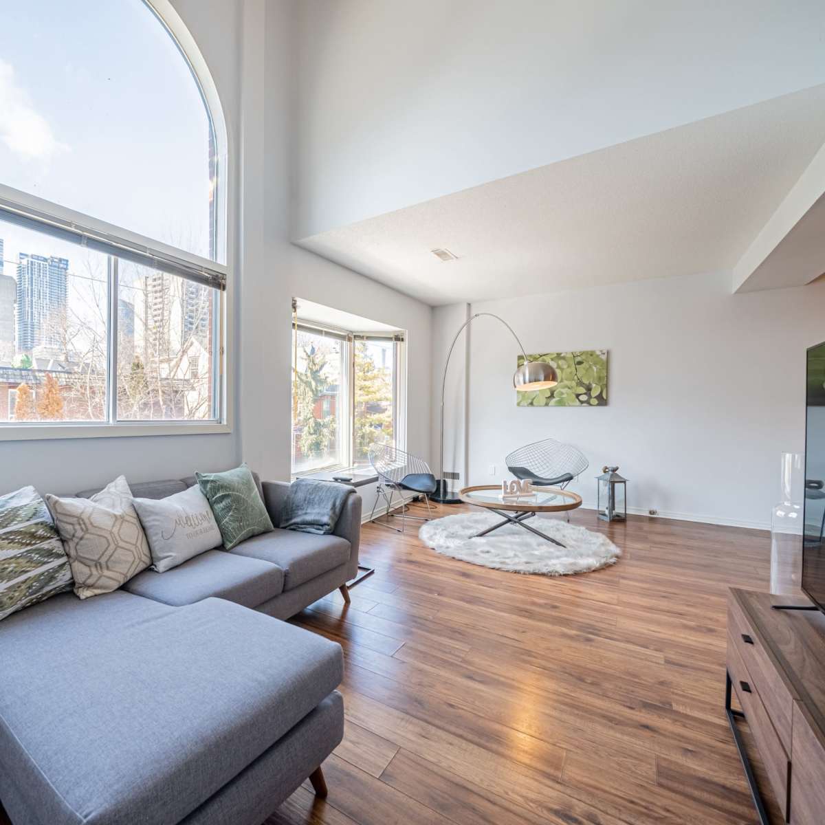 A modern living room features a gray sectional sofa, a round rug, and large windows letting in natural light, with city skyline views in the background.