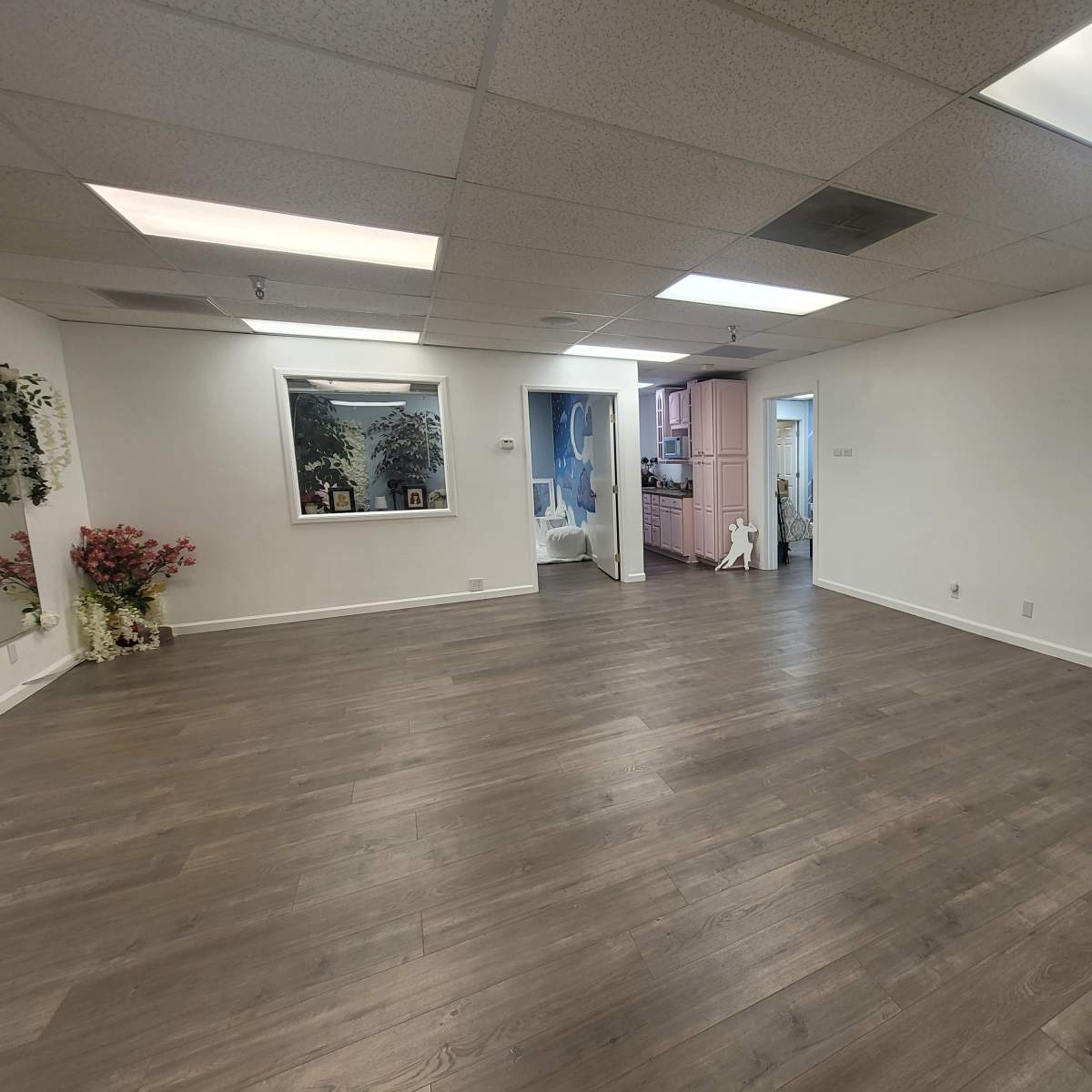 A spacious, empty room with wooden flooring, natural light from ceiling lights, and an open doorway leading to another area.