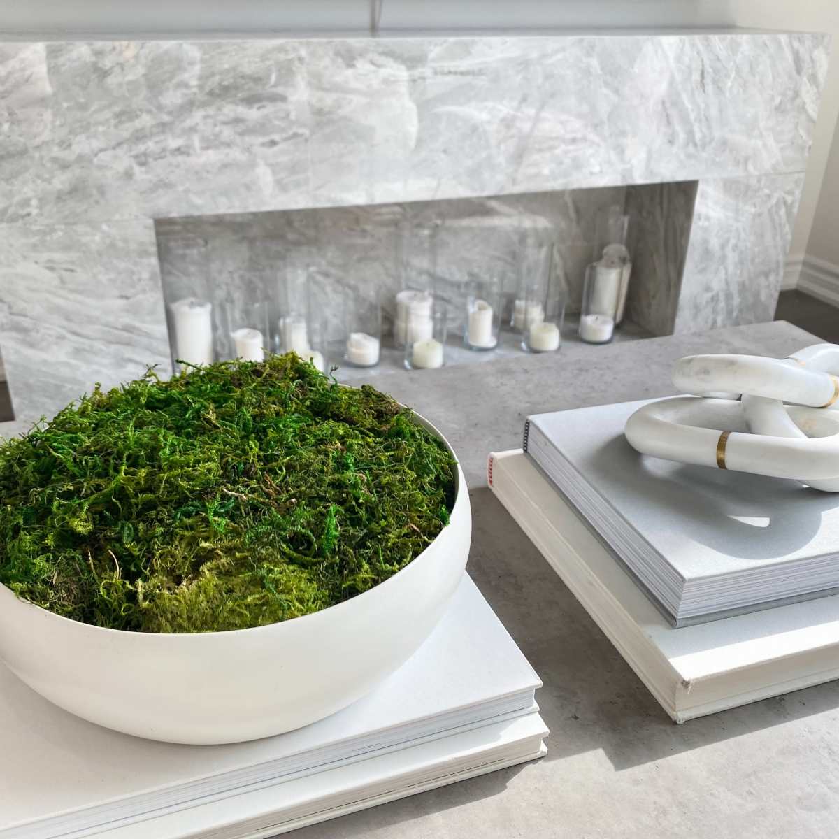 A white bowl filled with moss sits on a stack of gray books next to a marble fireplace with white candles arranged on the ledge.