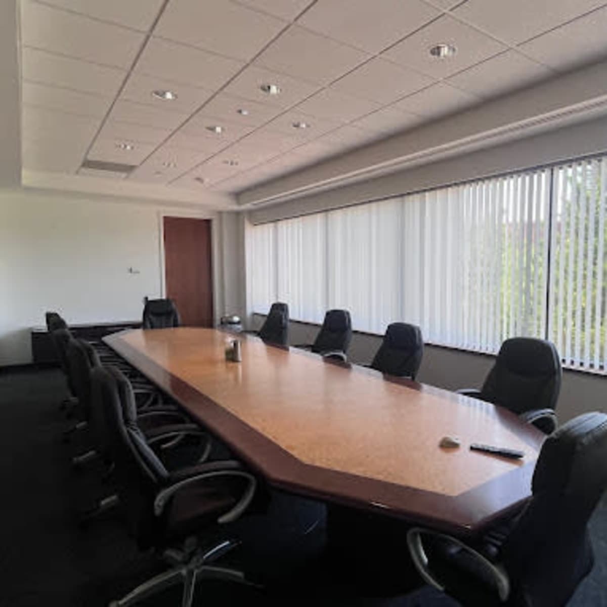 A conference room features a long table surrounded by black office chairs, with large windows allowing natural light to fill the space.