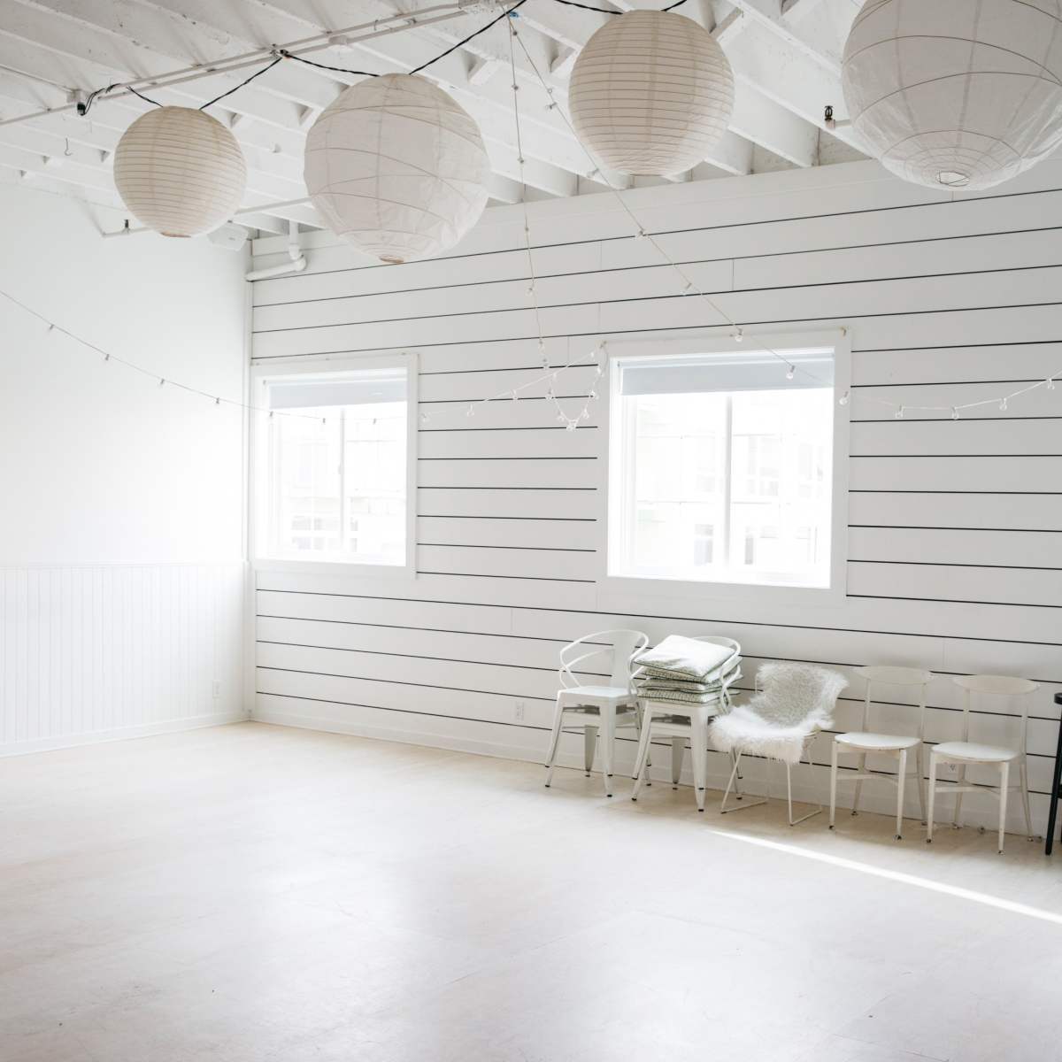 A bright, empty room features white walls with wooden paneling, overhead paper lanterns, and several chairs lined against the wall.