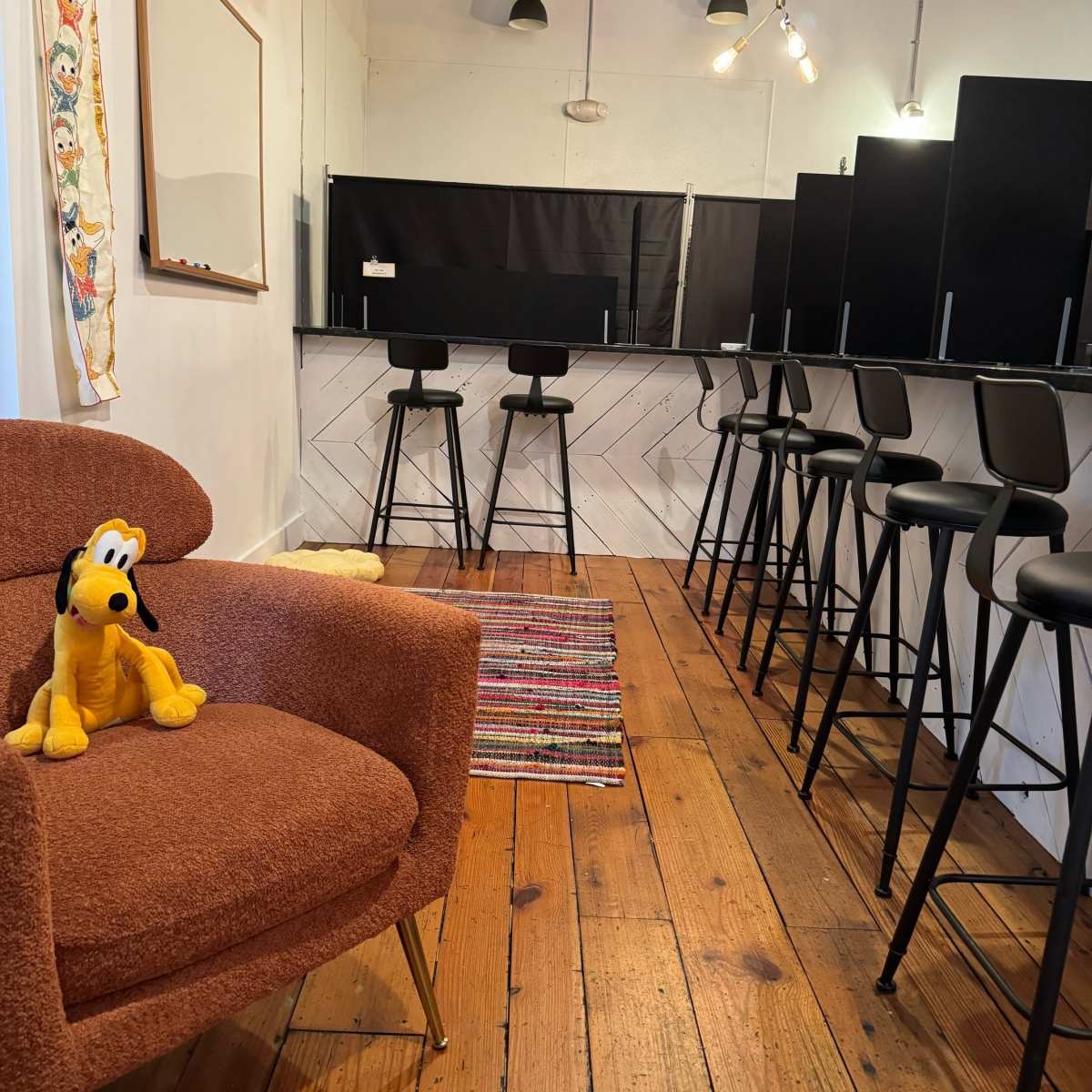 A spacious room with wooden floors, featuring a cozy brown chair and a bar area with black stools along a white wall.