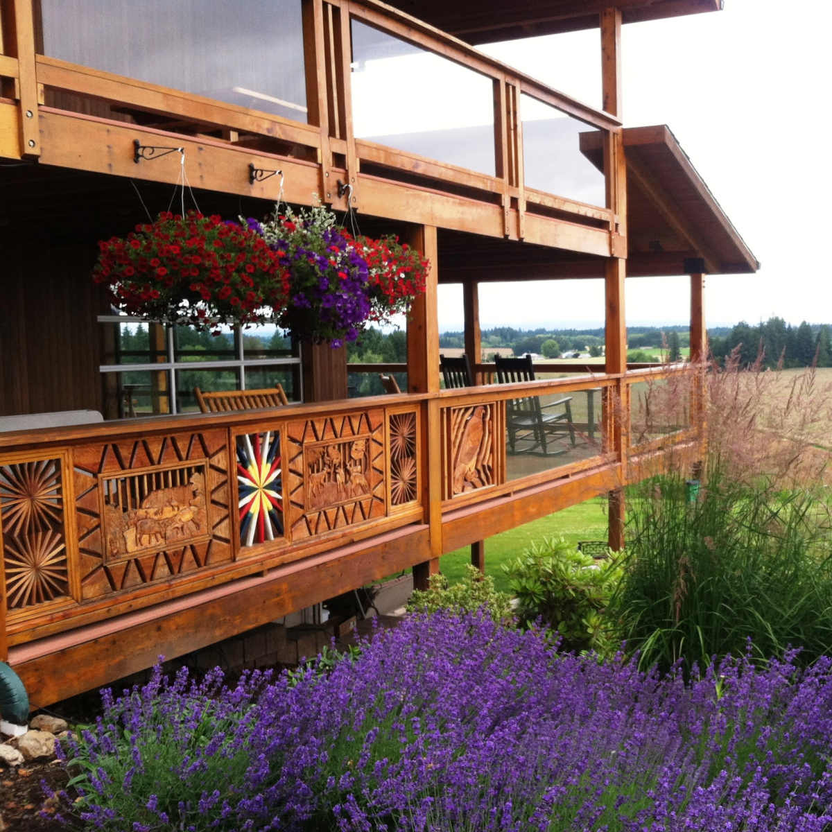 A wooden balcony with decorative railings overlooks a field, surrounded by purple flowers and hanging flower baskets.