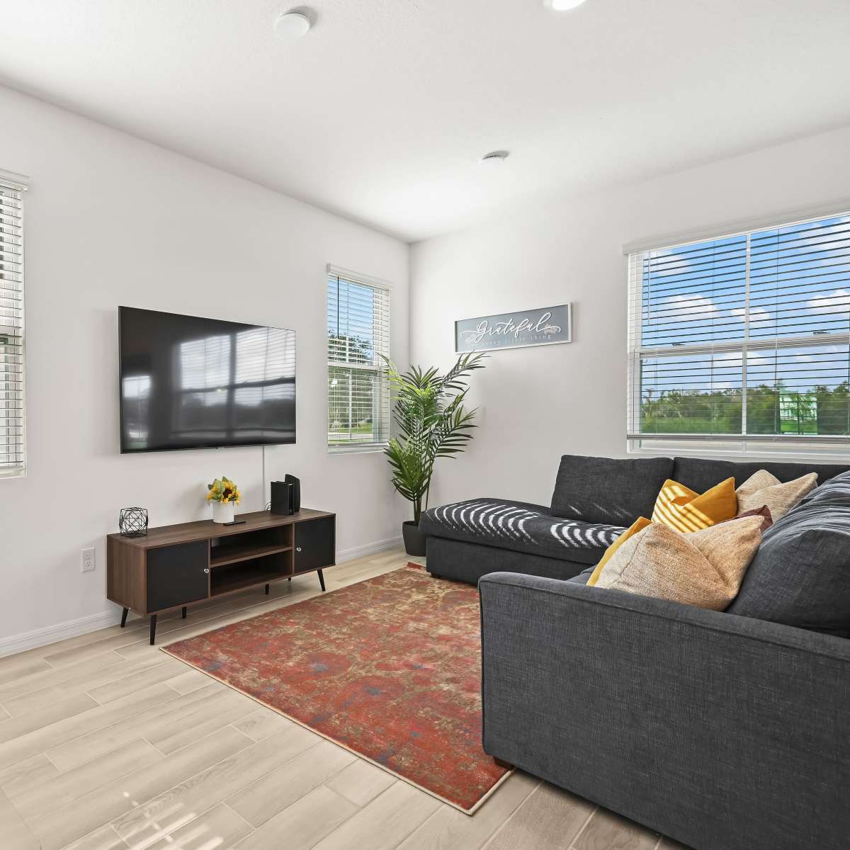 A modern living room features a sectional sofa, a wall-mounted television, a desk with a lamp, and a decorative rug.