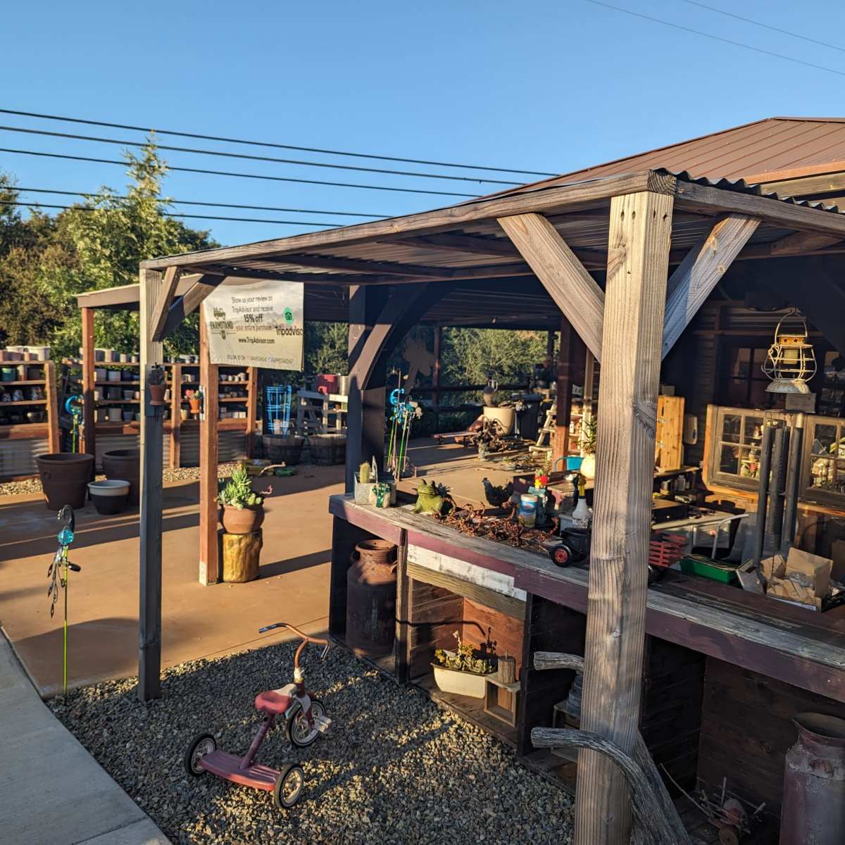 A rustic outdoor garden center with wooden structures displaying various gardening tools and pots, alongside a paved walkway and a few decorative items.
