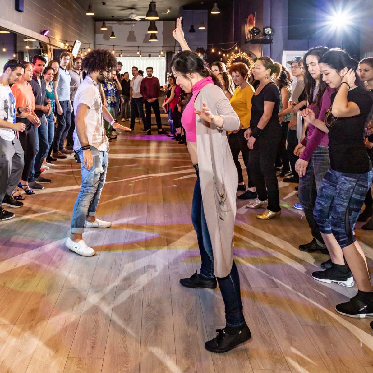 A large group of people are gathered in a dance studio, participating in a dance class with one instructor leading in the center.
