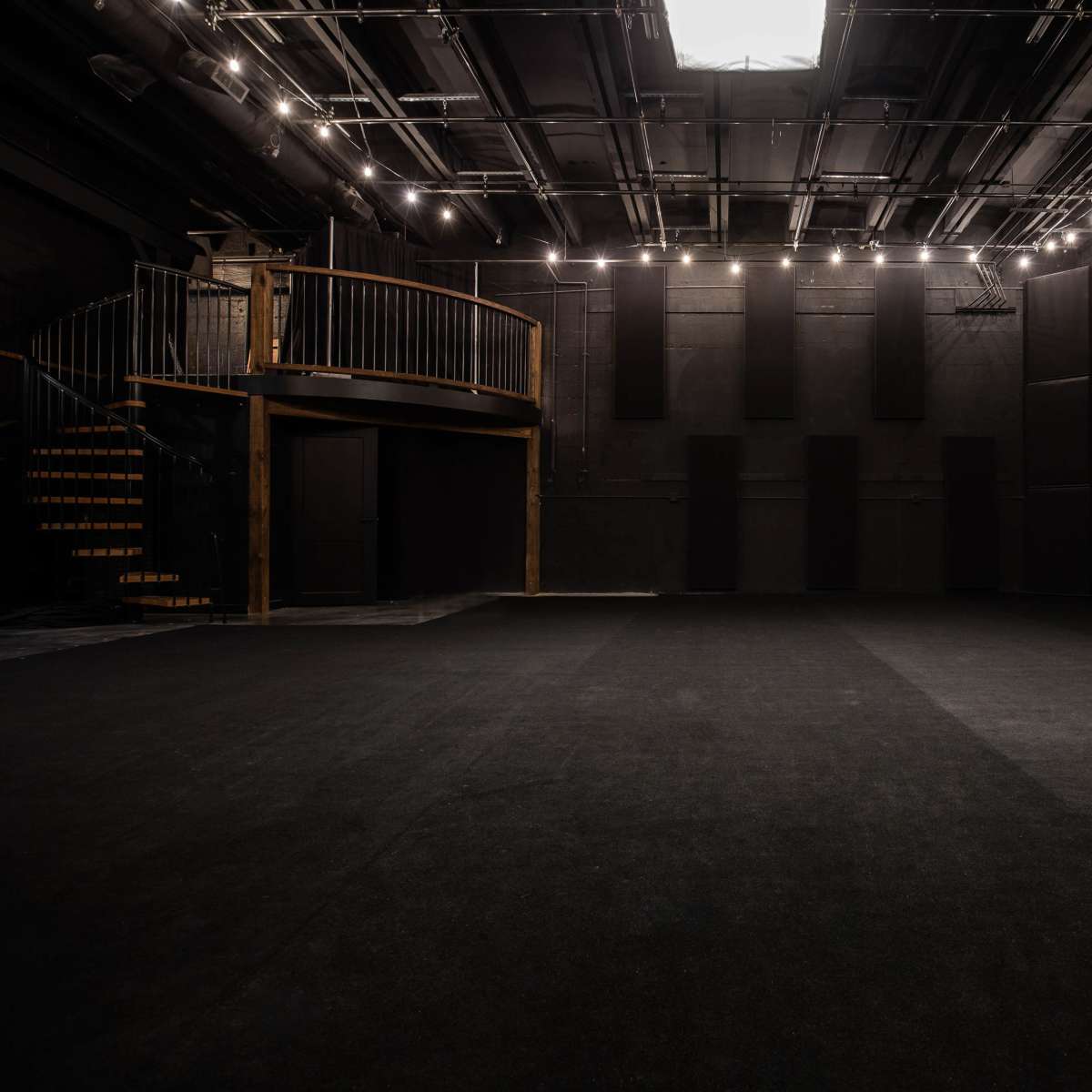 A dimly lit black box theater with a spacious, empty stage and a staircase leading to a balcony area.