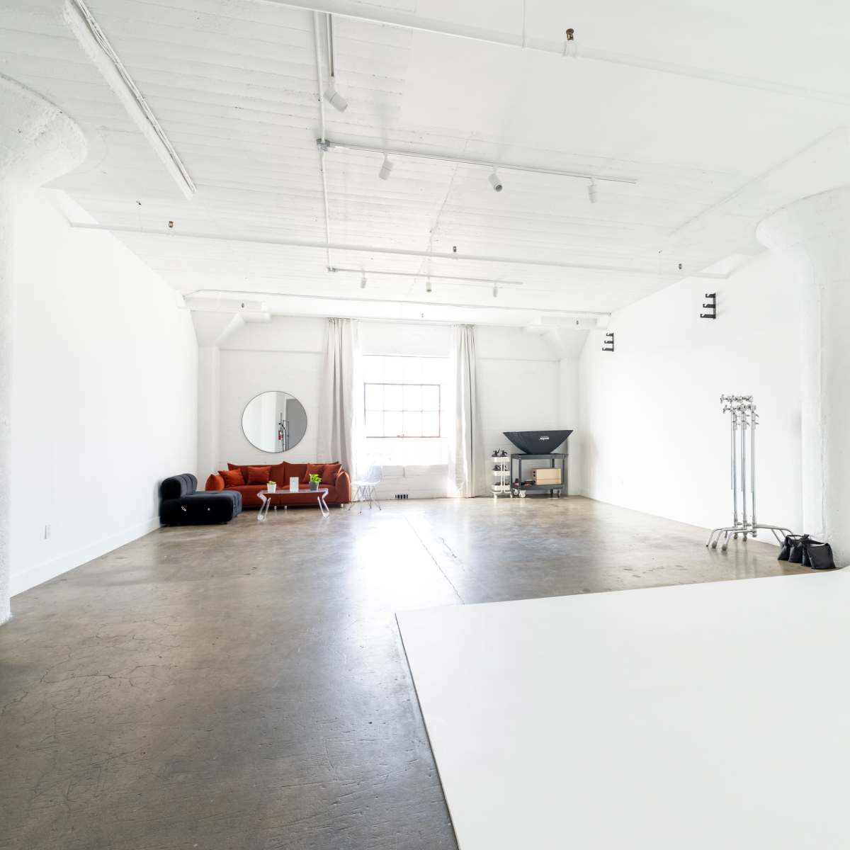 A spacious, empty studio with white walls, a concrete floor, a round mirror, and minimal furniture including a red couch and a television.