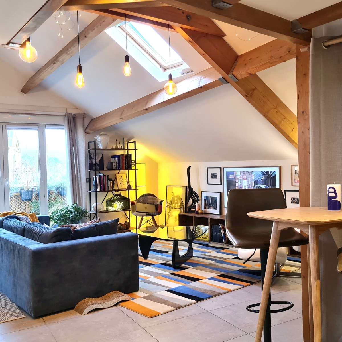 A modern living space with a high ceiling, featuring a sofa, a small table with stools, and a bookshelf illuminated by warm light.