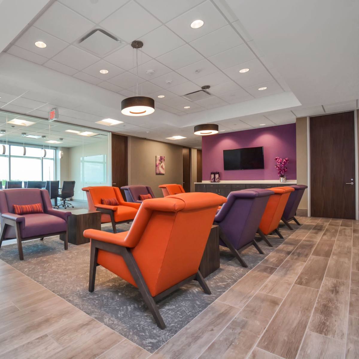 A modern office lounge area with orange and purple chairs, a conference room in the background, and large windows providing natural light.