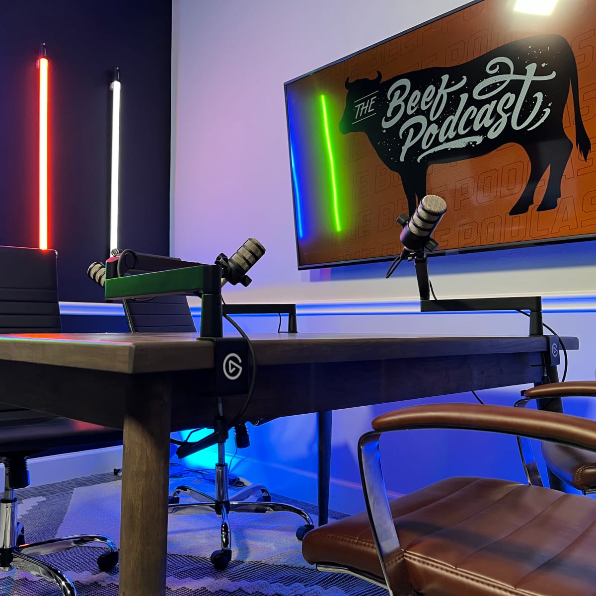 A podcast recording studio featuring a wooden table with microphones, chairs, and colorful LED light strips on the walls.