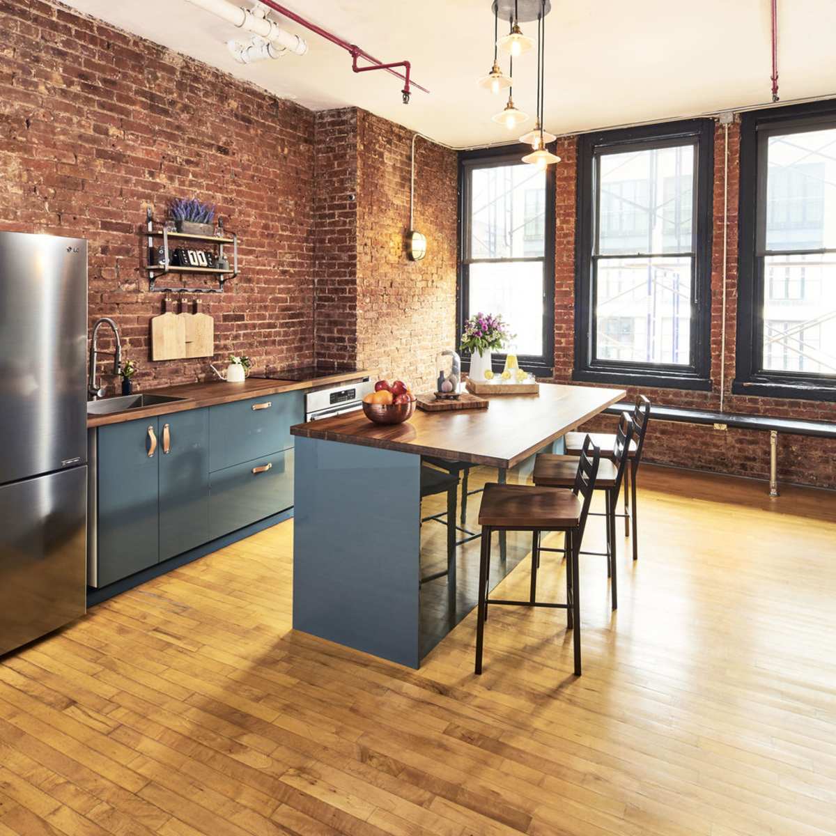 A modern kitchen with brick walls, a stainless steel refrigerator, a wooden island, and large windows that provide natural light.